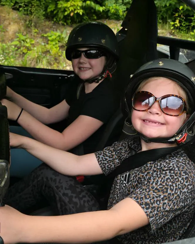 Two young riders in helmets and sunglasses smiling from the seat of a UTV