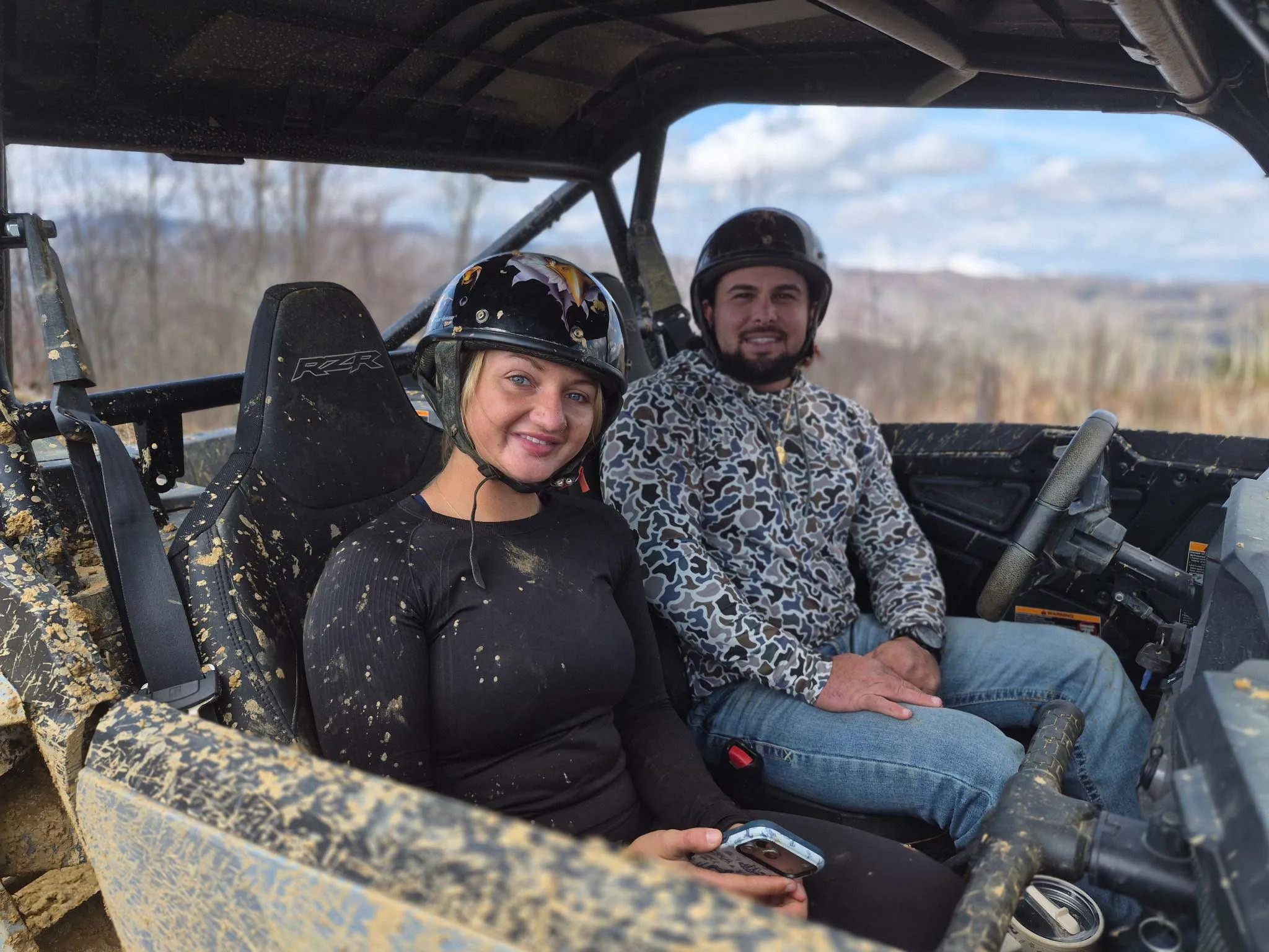 Two riders in helmets and goggles in the cab of a mud-covered UTV