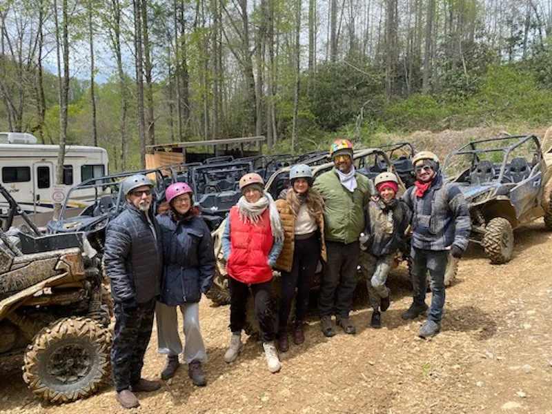 A large helmeted group beside the muddy UTVs in the forest