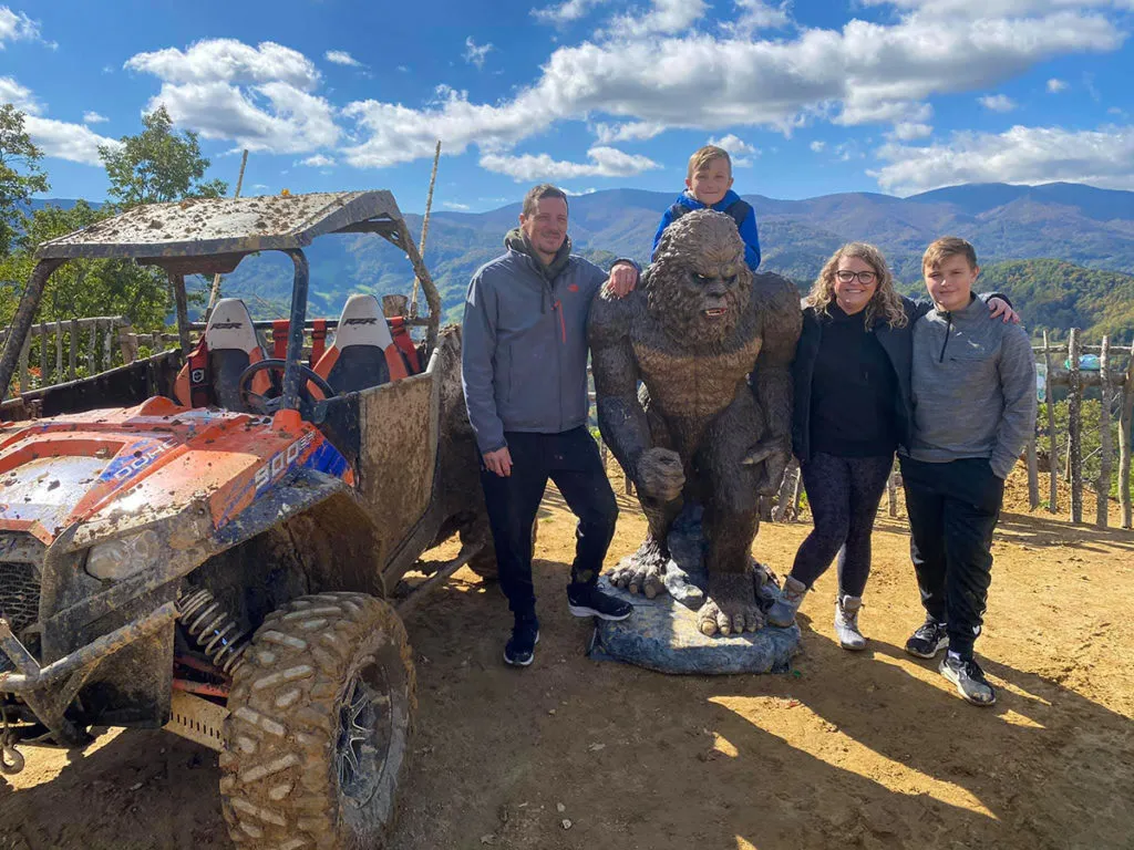 Five riders beside an orange UTV and a carved wooden bear sculpture at the summit overlook